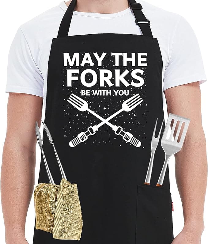 Black apron with “May The Forks Be With You” slogan worn by a grinning dad at the grill, pockets full of utensils and intergalactic confidence.